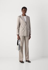 Helles grau gestreiftes Anzug mit einem maßgeschneiderten Blazer und Hosen. Das Modell hält eine silberne Tasche und trägt schwarze Ballerinas. Schlichte Accessoires vervollständigen den Look.