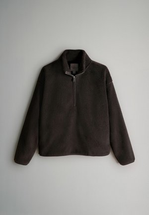 Brūns vilnas džemperis ar augstu apkakli un pusgaru rāvējslēdzēju. Raksturo mīksta, teksturēta virsma un brīvs piegriezums, ar gariem piedurknēm.