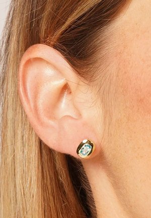 Gold stud earring with a light blue gemstone worn on the earlobe of a person with light brown hair.