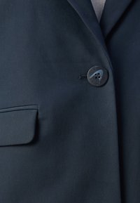 Navy blejzr s hladkým materiálem, zapínáním na jeden knoflík s názvem značky a náprsní kapsou s pečlivě sešitými okraji.