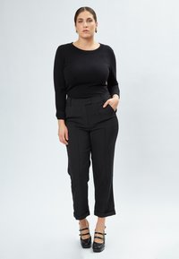 Schwarzes, eng anliegendes Langarmoberteil kombiniert mit hochgeschnittenen schwarzen Hosen, die eine flache Vorderseite und hochgekrempelte Ärmel haben, ergänzt durch schwarze Riemchen-Heels.