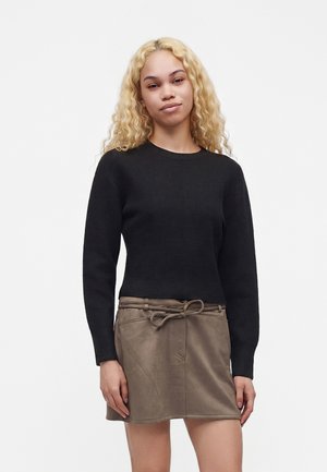Jeune femme aux longs cheveux blonds bouclés portant un pull noir à manches longues et une jupe marron avec une ceinture nouée.
