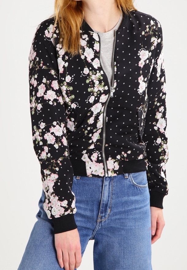 Personne portant une veste noire zippée avec un motif floral rose et blanc sur un haut gris et un jean bleu.