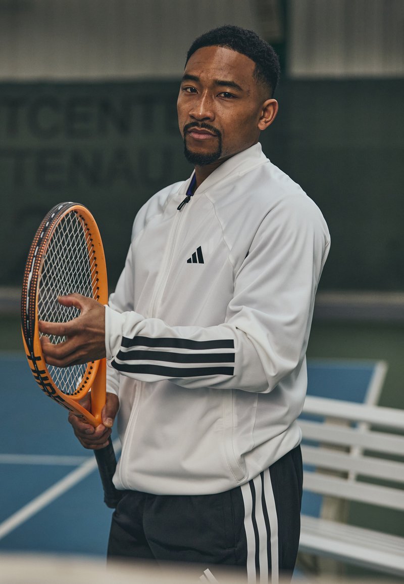 Witte zip-up jas met zwarte strepen, gedragen over bijpassende broek. Persoon houdt een oranje tennisracket vast en staat op een baan.