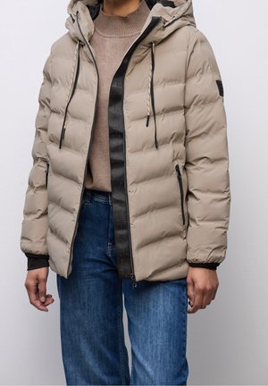 Osoba v béžové prošívané péřové bundě s kapucí, černým lemováním na zipu, hnědém svetru a modrých džínách na jednofarebném světlém pozadí.