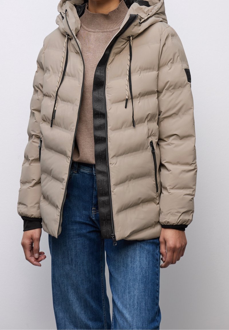 Personne portant une doudoune matelassée beige avec capuche, bordure zippée noire, pull beige et jean bleu sur un fond clair uni.