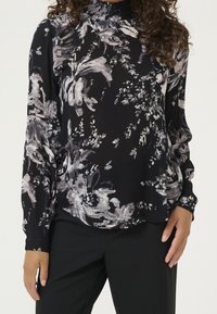 Blouse à manches longues en tissu noir avec des motifs floraux abstraits blancs et gris, dotée d'un col montant smocké et d'une coupe décontractée.