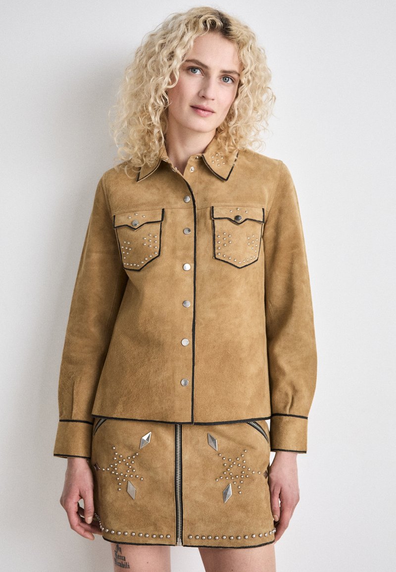 Camicia in camoscio tan con tasche anteriori, borchie argentate e piping nero; abbinata a una gonna coordinata con borchie geometriche e una cerniera.