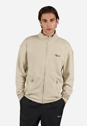 Beige zip-up jacket with ribbed texture, high collar, side pockets, and a subtle logo on the left chest, paired with black pants.