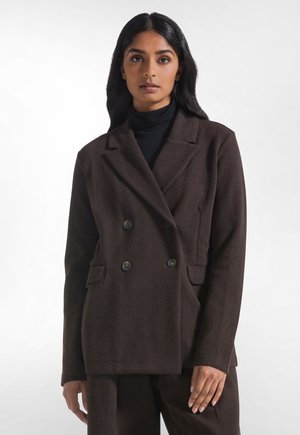 Donna con lunghi capelli neri indossa un blazer doppiopetto marrone scuro e pantaloni abbinati sopra un dolcevita nero, su uno sfondo semplice.