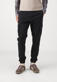Pantalones cargo negros con tobillos ajustados, que cuentan con grandes bolsillos laterales, un tejido duradero y detalles de costura visibles. Combinados con zapatillas marrones.