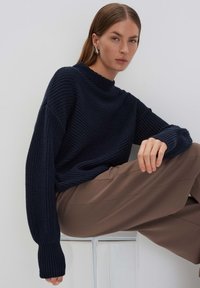 Maglione lavorato a maglia blu navy con collo alto, maniche lunghe e texture a costine. Abbinato a pantaloni beige e posizionato su uno sfondo chiaro.