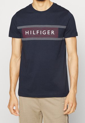 Man draagt een marineblauw shirt met korte mouwen met "HILFIGER" in witte letters op een rood-wit gestreept rechthoekig ontwerp over de borst.