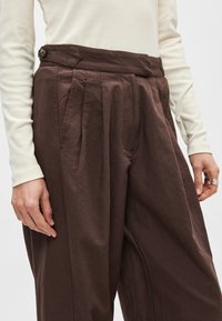 Καφέ, πιετέ παντελόνια με ψηλή μέση, με λεπτομέρεια κουμπιού στη μέση και πλαϊνές τσέπες, κατασκευασμένα από υφασμάτινη υφή.