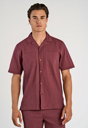 Chemise à manches courtes en tissu maroon texturé, avec un col camp, dotée de deux poches avant et de boutons assortis.