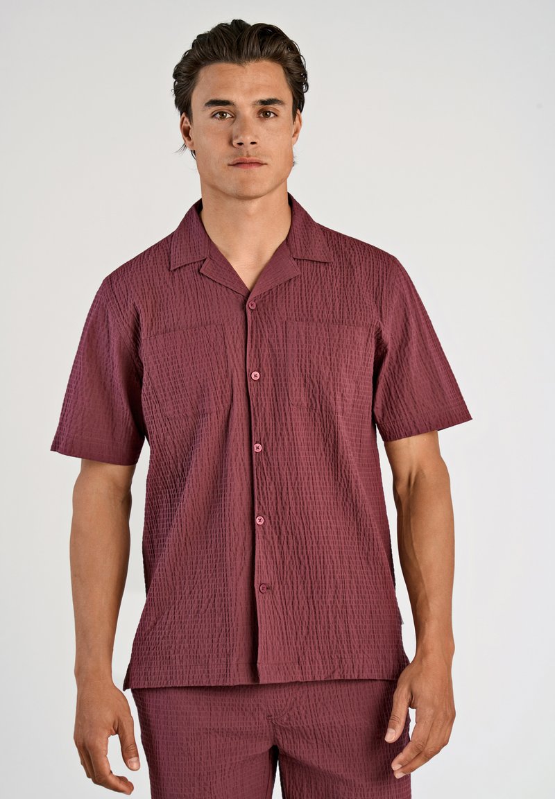 Camicia a maniche corte in un tessuto borgogna testurizzato con colletto a campagna, dotata di due tasche frontali e bottoni coordinati.