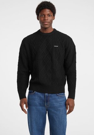 Maglione nero lavorato a maglia con motivi a rombo testurizzati, scollo rotondo, e polsini e orlo a costine, indossato con jeans blu.