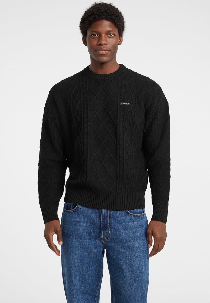 Maglione nero lavorato a maglia con motivi a rombo testurizzati, scollo rotondo, e polsini e orlo a costine, indossato con jeans blu.