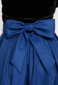 Sininen satiinimekko, jossa on suuri solmu vyötäröllä, pleateilla solmun alapuolella ja musta samettinen yläosa. Yhteensä sileä rakenne.