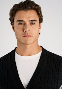 Czarny dziergany kardigan z warkoczowym wzorem, szerokim dekoltem w serek, noszony na białej koszulce z okrągłym dekoltem. Gładka faktura, dopasowany krój.