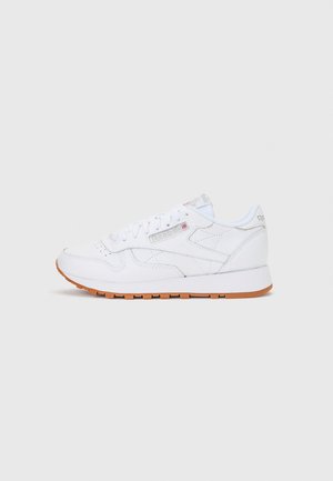 Biały skórzany sneaker Reebok z gumową podeszwą, perforowanymi detalami oraz firmowym logo po boku, na języku i pięcie na jednolitym tle.