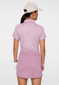 Donna che indossa un completo da golf lilla chiaro, guanto bianco e cappellino rosa chiaro, vista da dietro su uno sfondo bianco semplice.