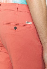 Pantaloni Wrangler color corallo con una texture liscia, dotati di una tasca posteriore con chiusura a bottone e un'etichetta con marchio.