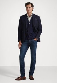 Ναυτικό σακάκι πάνω από γιλέκο σε ναυτικό μπλε, λευκό ριγέ πουκάμισο και σκούρα μπλε τζιν. Καφέ μοκασίνια συμπληρώνουν την εμφάνιση. Το σακάκι έχει χρυσά κουμπιά.