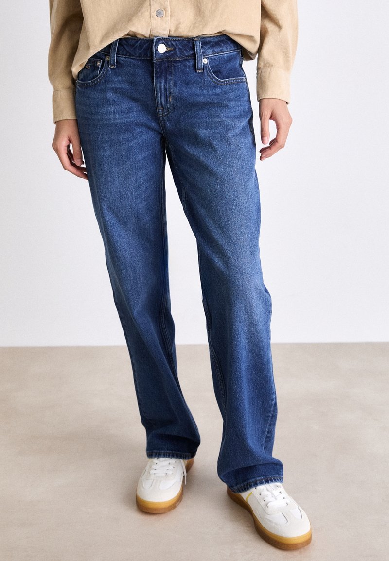Dark blue denim jeans with a straight leg fit, five-pocket design, and a button closure, paired with white sneakers.