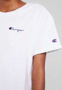 Biała bawełniana koszulka z krótkim rękawem, z niebieskim haftowanym logo "Champion" na piersi oraz małym logo na rękawie.