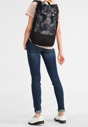 Henkilö pukeutuneena istuviin sinisiin farkkuihin, valkoisiin kenkiin, vaaleanpunaiseen verkkopaitaan ja musta-harmaaseen, soljilla varustettuun reppuun.