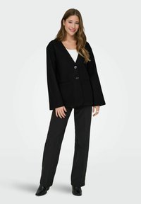 Veste noire oversize avec un col en V, deux boutons et des poches latérales, associée à un pantalon noir et des bottines. Texture de tissu lisse.