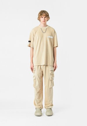 Camiseta oversized de rayas beige con un bolsillo frontal, combinada con pantalones cargo a juego que cuentan con múltiples bolsillos laterales. Se lleva con zapatos texturizados beige.