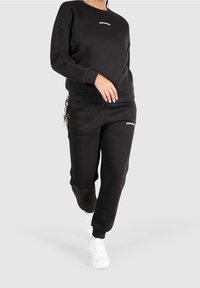 Svart bomullströja och joggers med rund hals och logotyp på bröstet och låret, tillsammans med vita träningsskor.