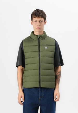 Weicher olivgrüner Weste mit hohem Kragen, Reißverschluss vorne und Logopatch. Kombiniert mit einem schwarzen kurzärmligen Hemd und blauen Jeans.
