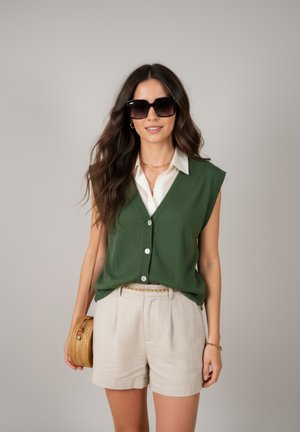 Vrouw met lang bruin haar die zwarte zonnebril draagt, een groene mouwloze vest met knopen, een wit overhemd, beige korte broek en een ronde strooien tas bij zich heeft.