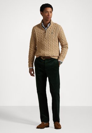Sweater beige en tricot avec une fermeture éclair quart, pantalon en velours côtelé vert et chaussures en daim marron. Présente une chemise avec des rayures blanches et bleues en dessous.