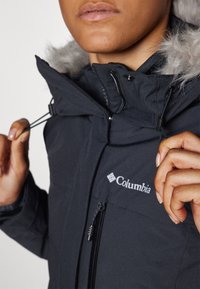 Chaqueta aislante negra con capucha forrada de piel sintética, cordones ajustables y un bolsillo delantero con cremallera. Logo de Columbia visible en el pecho.