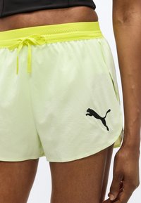 Κοντινό πλάνο ατόμου που φοράει αθλητικό σορτς Puma σε ανοιχτό κίτρινο χρώμα με σκοινί στη μέση και μαύρο λογότυπο Puma στο αριστερό πόδι.