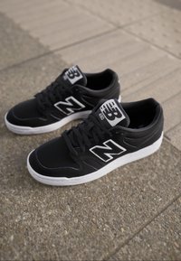 Czarne skórzane sneakersy z białymi akcentami, z wyraźnym logo "N", okrągłym noskiem i sznurowanym przodem. Podeszwa jest w przeważającej części biała.