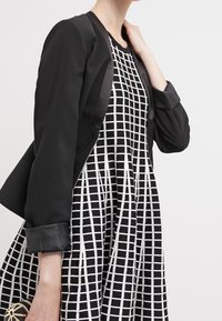 Femme portant une robe sans manches à motif quadrillé noir et blanc avec une veste noire posée sur les épaules, sur un fond uni.