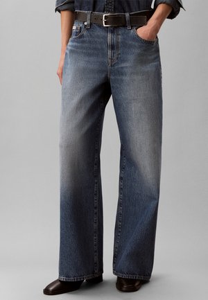 Person iført vidde falmede blå jeans, sort bælte, mørke sko, med den ene hånd i lommen mod en ensfarvet grå baggrund.