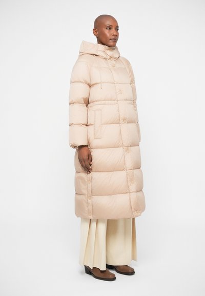 Töltött bézs kabát kapucnival, gombos elülső résszel, nagy zsebekkel és térd alá érő fazonnal. A modellen krém színű bőszárú nadrág és barna cipő van.