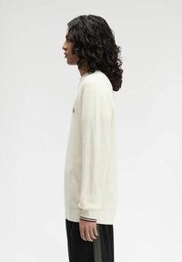 Cremfarbener gestrickter Pullover mit einem strukturierten Muster, gerippten Bündchen und einem kleinen gestickten Logo auf der Brust, getragen über dunklen Hosen.