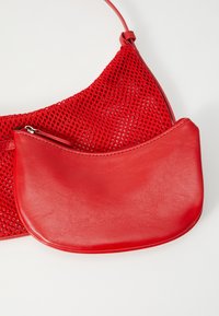 Sac à main rouge avec un design en filet et une poche avant en cuir lisse, fermée par une fermeture éclair. Bandoulière ajustable incluse.