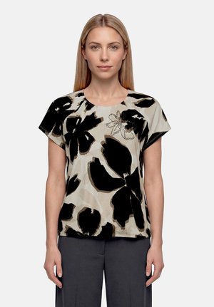 Femme aux cheveux blonds et raides porte un chemisier beige à manches courtes avec un grand imprimé floral noir et un pantalon gris, debout devant un fond Uni.