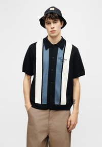 Νεαρός άνδρας που φοράει μαύρο καπέλο τύπου bucket, μαύρο κοντομάνικο πουκάμισο με μπλε και λευκές κάθετες ρίγες, και μπεζ παντελόνι, στέκεται με τα χέρια στις τσέπες.