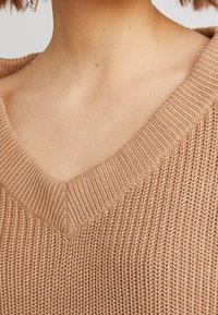 V-neck knitted sweater in a light brown color with a textured, ribbed design and a wide neckline.