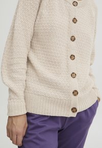 Beige kötött kardigán texturált mintával, gombos elülső résszel és bordás mandzsettákkal. Lila, lágyan formázott nadrággal párosítva.