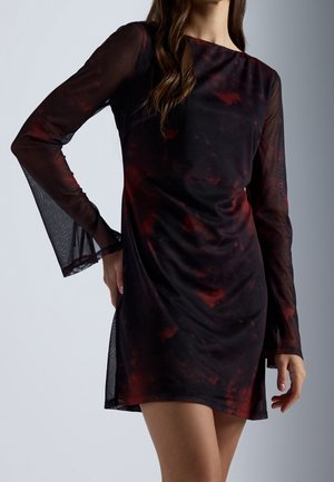 Femme portant une robe mini noire et rouge foncé à motif, transparente, manches longues et silhouette ajustée, sur un fond clair uni.